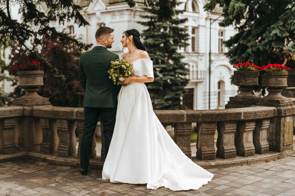 Cuplu îmbrățișat în ziua nunții, fotografie în grădină, stil film look, Enny Alex – fotograf nuntă București Mire și mireasă în ținute elegante, fotografie cinematică de nuntă în natură – București Fotografie romantică de nuntă, stil analog, realizată de Enny Alex în grădină